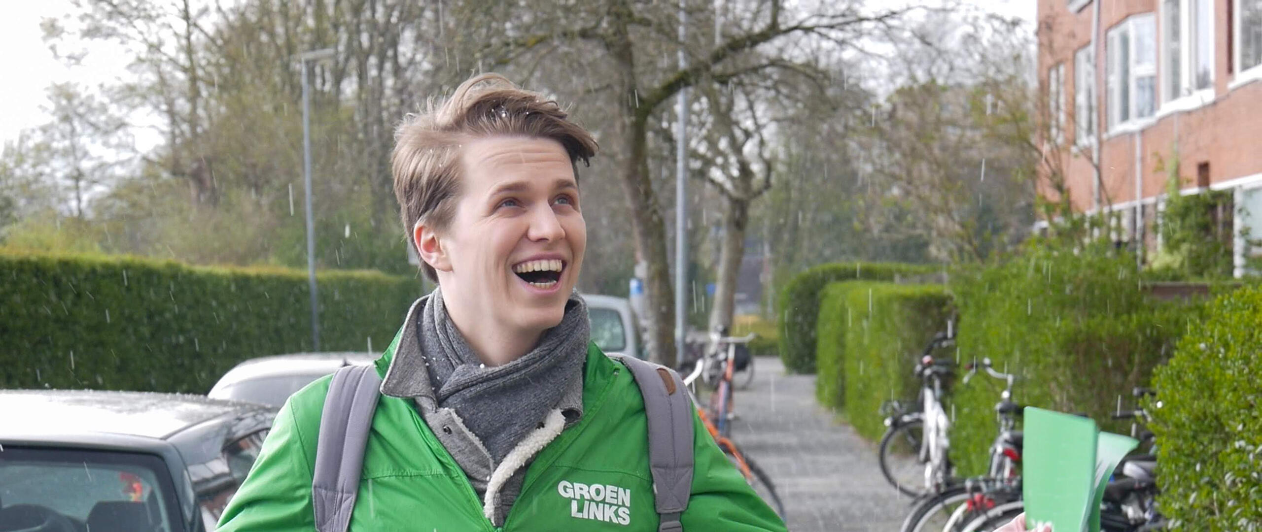 Een GroenLinks-vrijwilliger kijkt blij terwijl hij in de regen flyers vasthoudt